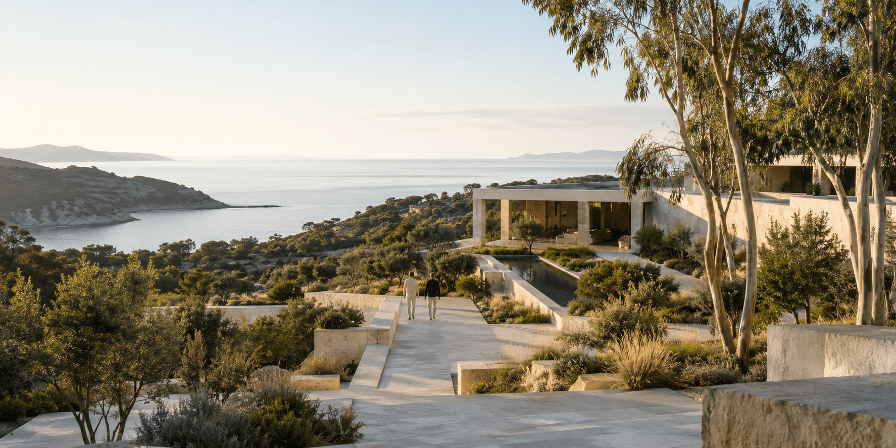 Two people walking a stone pathway toward a modern hillside residence overlooking the coastline at sunset.